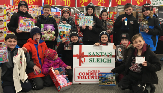 A group of young children holding up gifts purchased for the Santa's Sleigh Christmas Program. In the center of the picture, two boys are holding a Santa's Sleigh poster 
