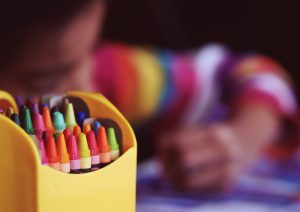 A box of colorful crayons at the front left hand side of the picture. A young child is blurred in the background coloring.