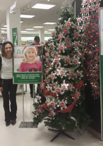 A woman stands next to a Tree of Wishes sign depicting a smiling little girl. The sign is next to a Christmas tree decorated with red ornaments.