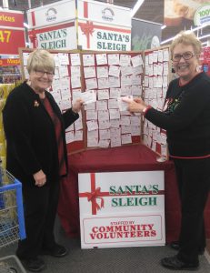 Two woman standing in front of the give-a-gift board for the Santa's Sleigh Christmas program, which is covered in the names of Children in the care of the CAS