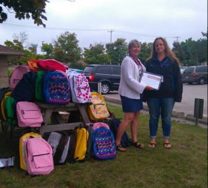 Two foundation workers stand next to a picnic table covered with backpacks that have been donated to the Backpacks For Kids program.