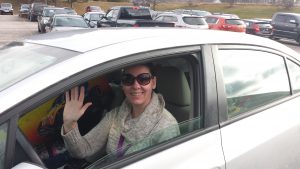 A woman wearing sunglasses sitting in the driver's seat of a car. She is smiling and waving.