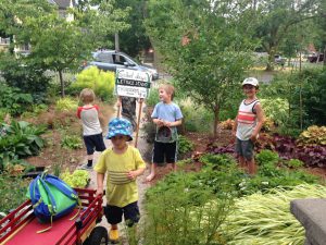Four kids stand outside in an area with lots of trees and greenery, near a Salad Days sign. One of them is pushing a red wagon with a backpack inside.