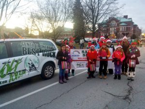 A group of children wearing red festive clothing and Santa hats, standing beside a Wolf 101.5 car.