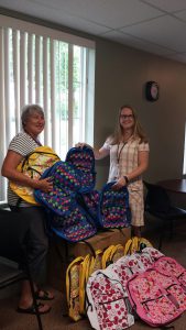 Two foundation members holding several backpacks that have been donated, near a small pile of more backpacks.