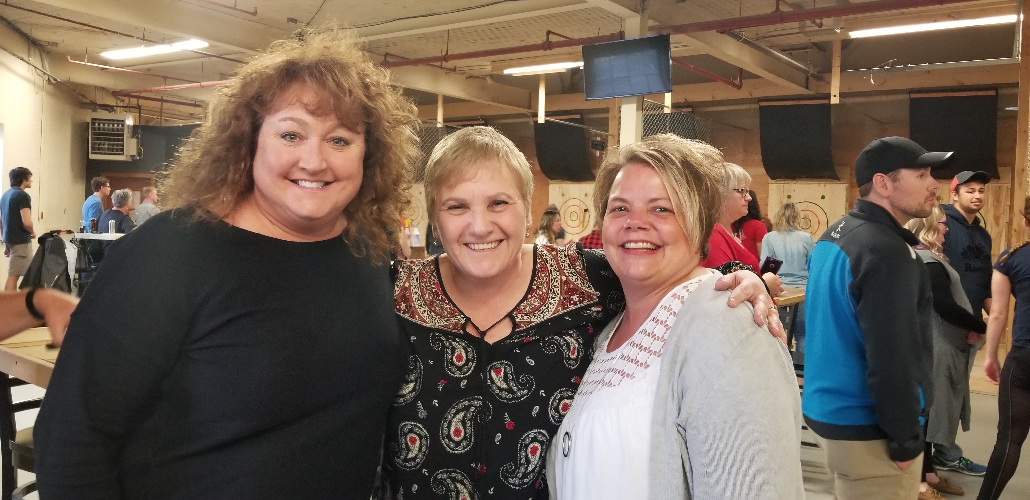 Three women in the Axe Club building, facing the camera and smiling. The one standing in the middle has her arms around the others' shoulders.