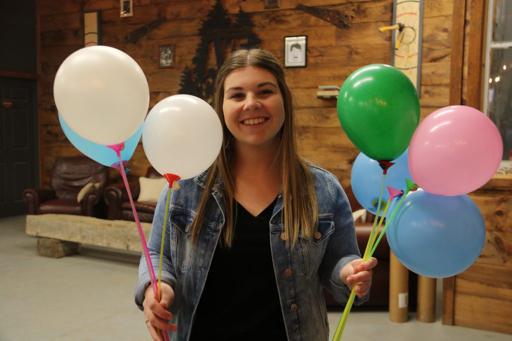 A woman holding several blue, white, green, and pink balloons in each hand, smiling.
