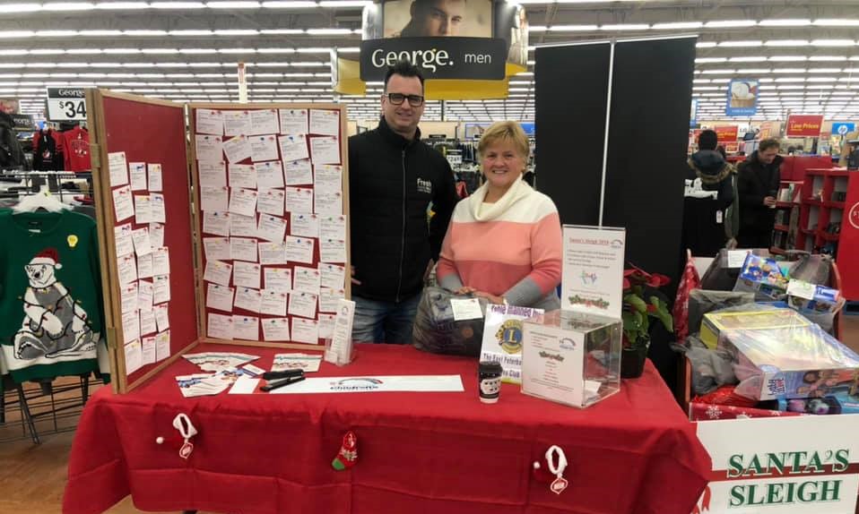 Two volunteers standing to the right of the give-a-gift board at Walmart, during the Santa's Sleigh Christmas program.