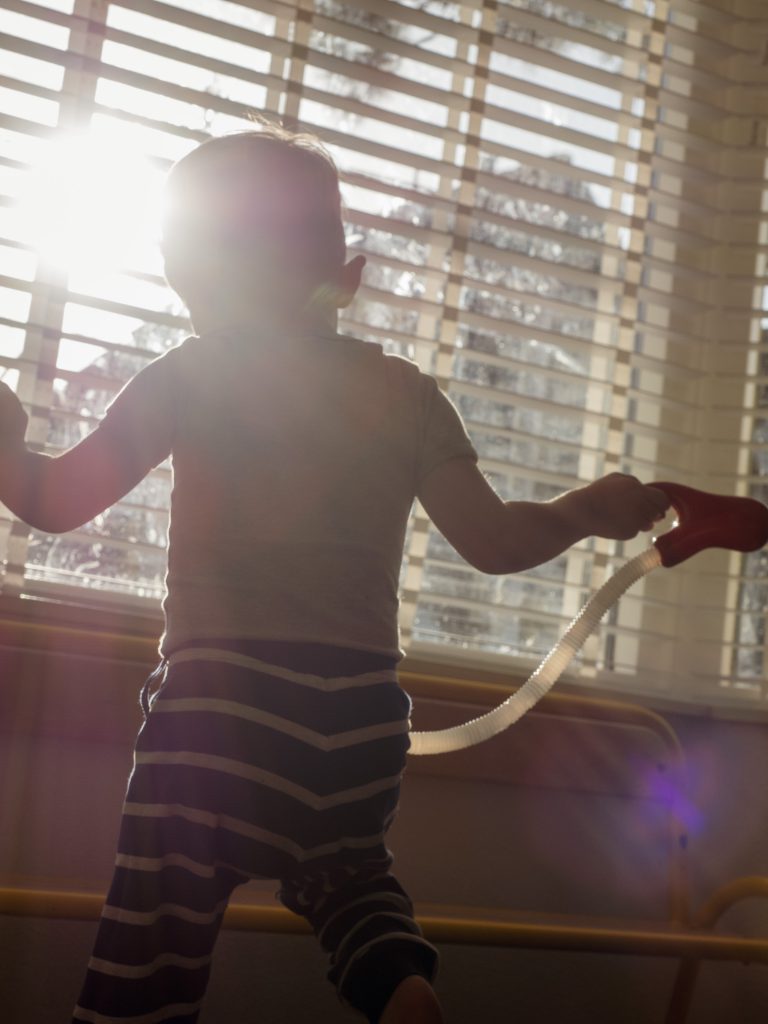 A little boy stands in front of a window with its blinds open, the sun shining through.
