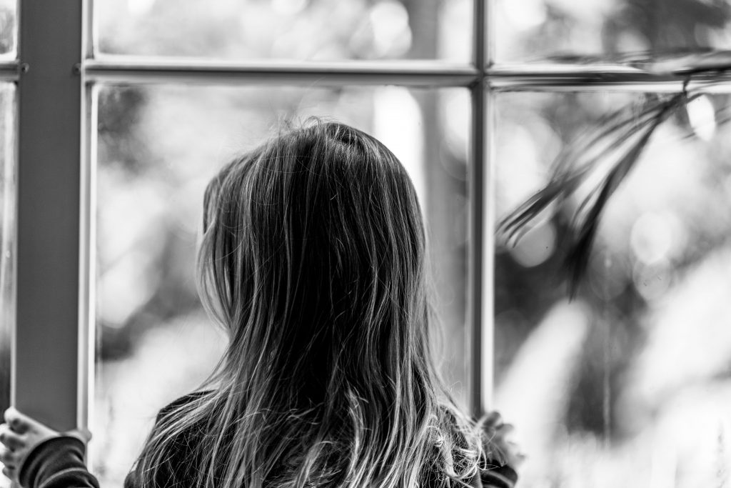 A greyscale photo of a little girl with her back to the camera, looking out of a window.