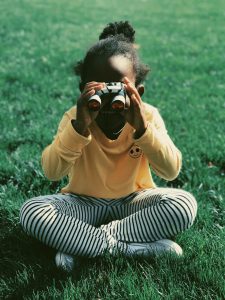 A little girl sitting on grass, looking through a pair of binoculars.