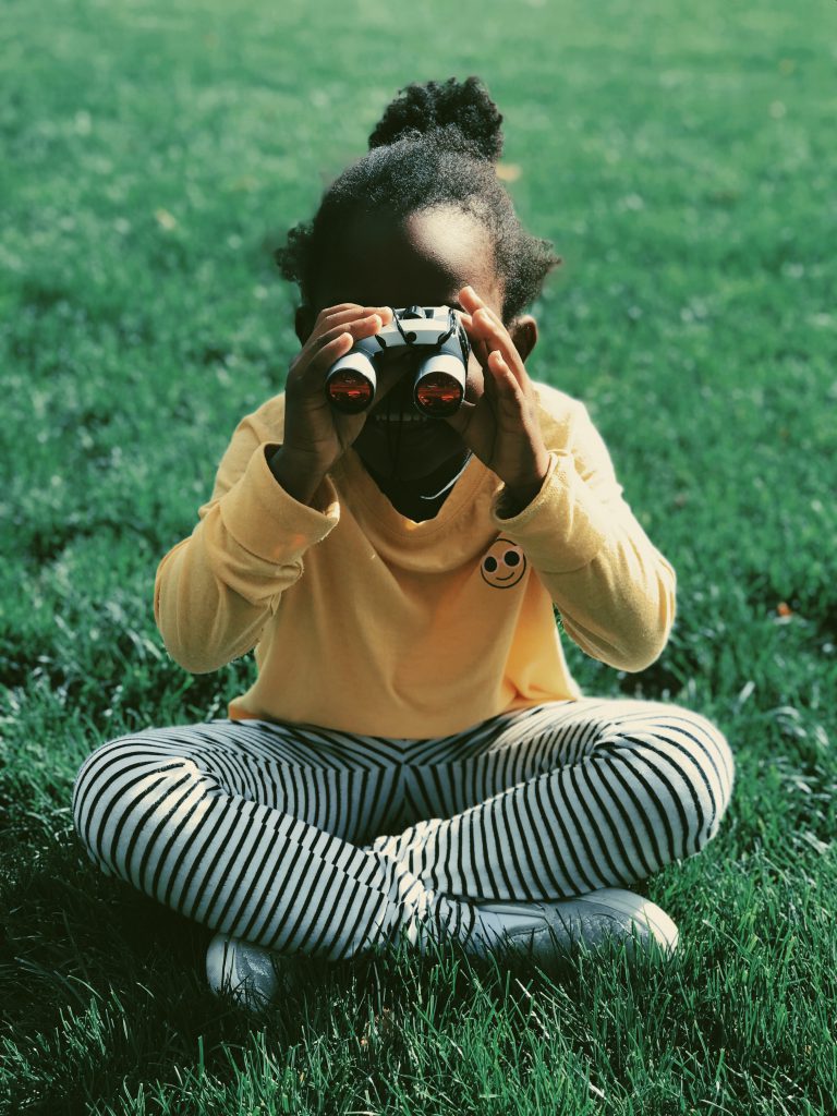 A little girl sitting outdoors, looking through a pair of binoculars.