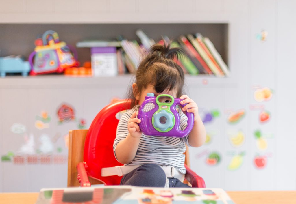 A toddler sitting in a high chair takes a photo using a purple toy camera.
