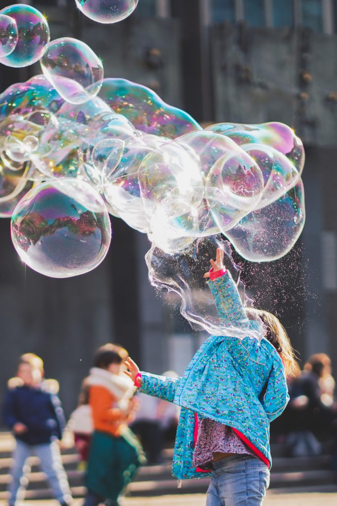 A child in a blue coat reaches out to pop a large amount of bubbles that are floating towards them.