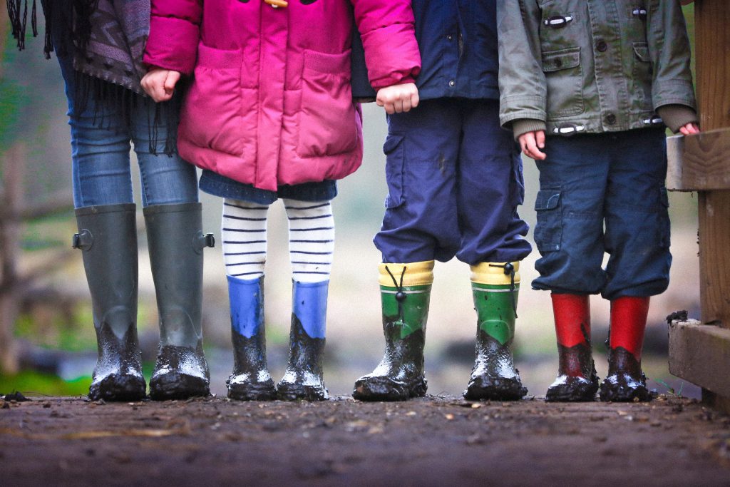 Four kids standing side by side facing the camera, visible from the chest down. They are wearing different-coloured coats and rainboots.