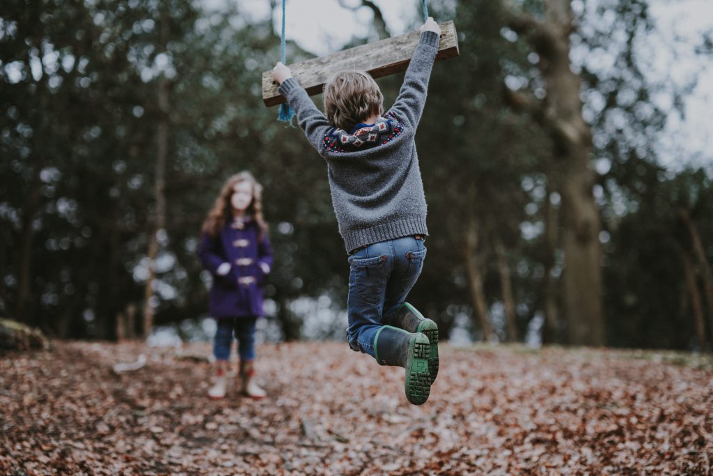 A child holding onto a wooden swing, while an older woman watches.