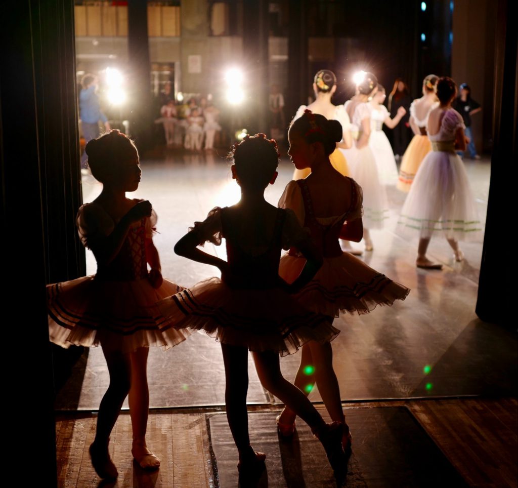 Three little girls in ballerina outfits wait backstage while another group of dancers leave the stage. They are dimly lit.