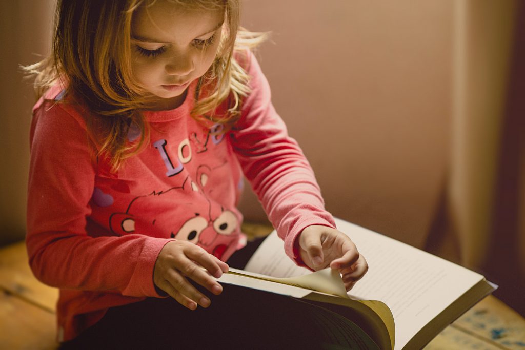 A little girl in a pink shirt reads a large book.