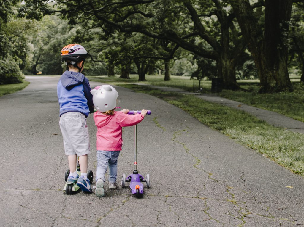 Two children on an outdoor path; one is riding a three-wheeled scooter, while the other is walking with the scooter beside her.