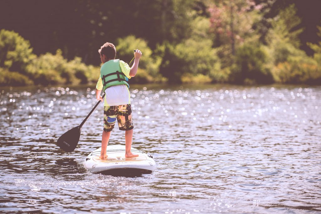 A child wearing a lifejacket, rowing a paddleboard through a lake.