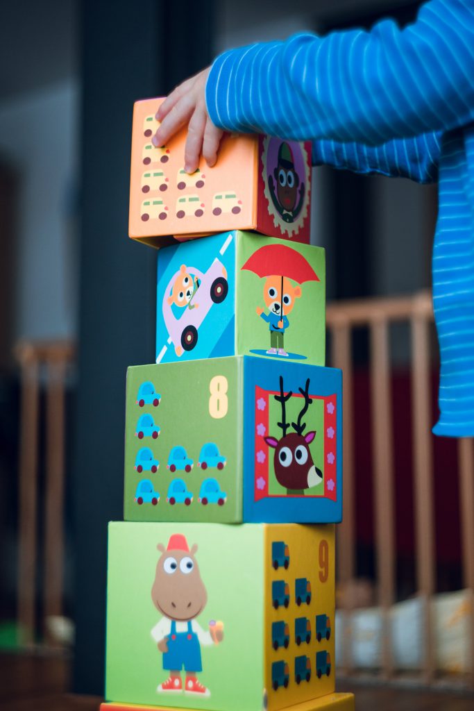 Four multicoloured building blocks stacked on top of one another; the topmost one is being placed there by a young child.