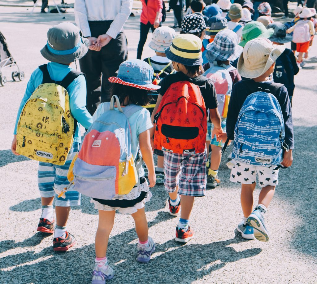 Four children with colourful backpacks walk down the street together, facing away from the camera.