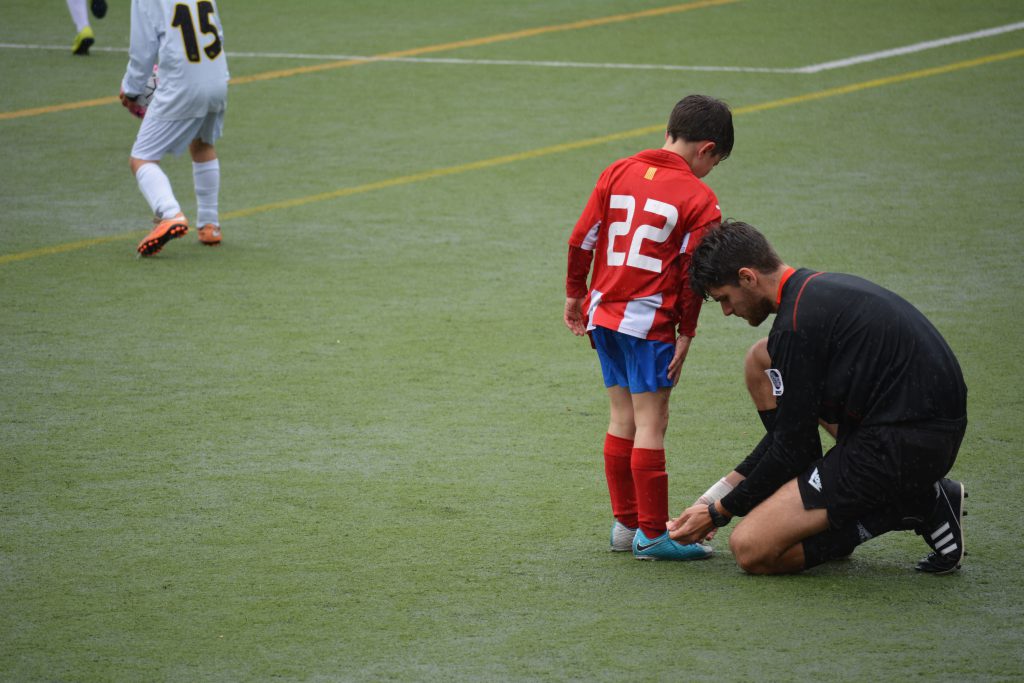 A child wearing a soccer uniform and standing on a soccer field, while a coach ties his shoe,