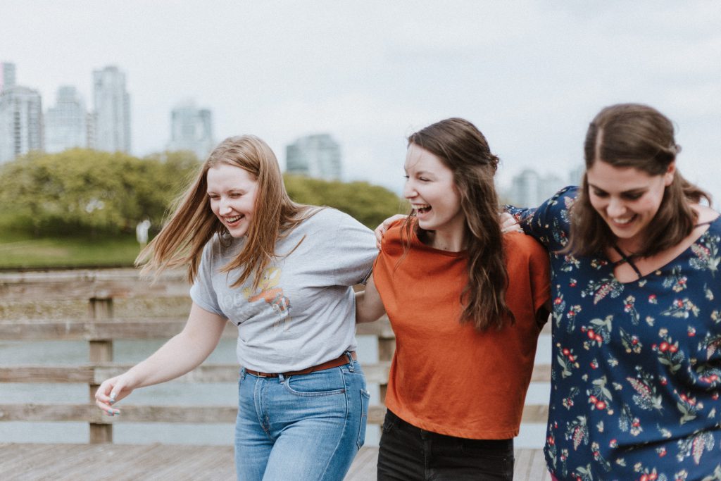 Three teenage girls with their arms around each other's shoulders, laughing and walking together on a wooden bridge.