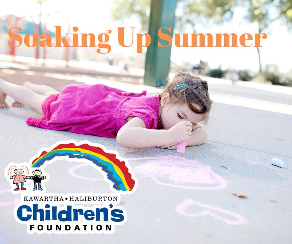 A little girl lying on her stomach on the sidewalk, drawing a heart with chalk. The text above her reads Soaking Up Summer. The KHCF logo is in the bottom left corner.