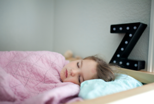 A child sleeps in a bed, covered with a pink blanket. A large decorative letter Z rests on the bedframe.