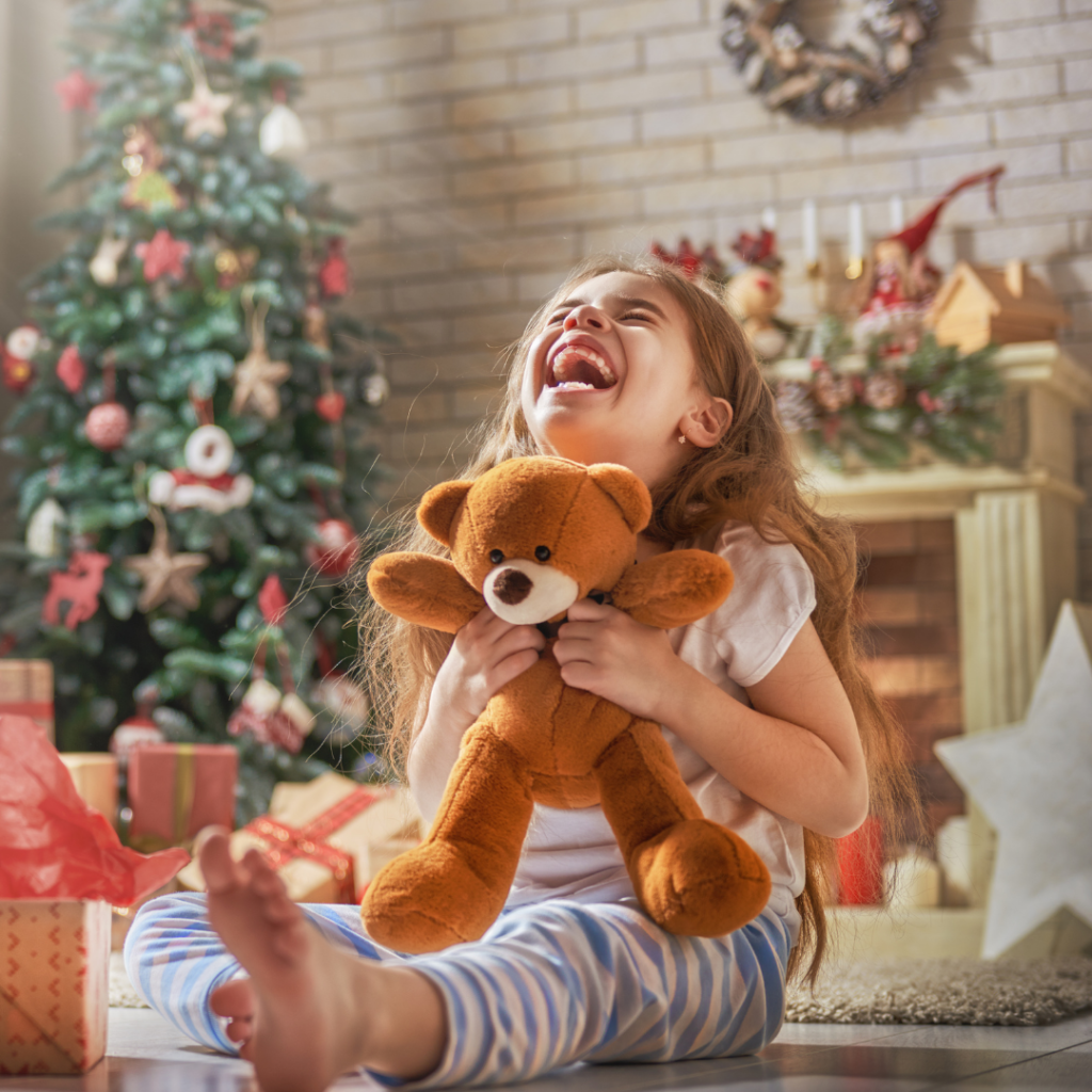 A child sits near a Christmas tree, holding a teddy bear and laughing.